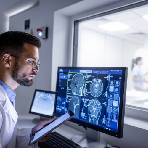 Doctor examining X-ray images on display in control room while in background nurse preparing the patient for new MRI scanning test.